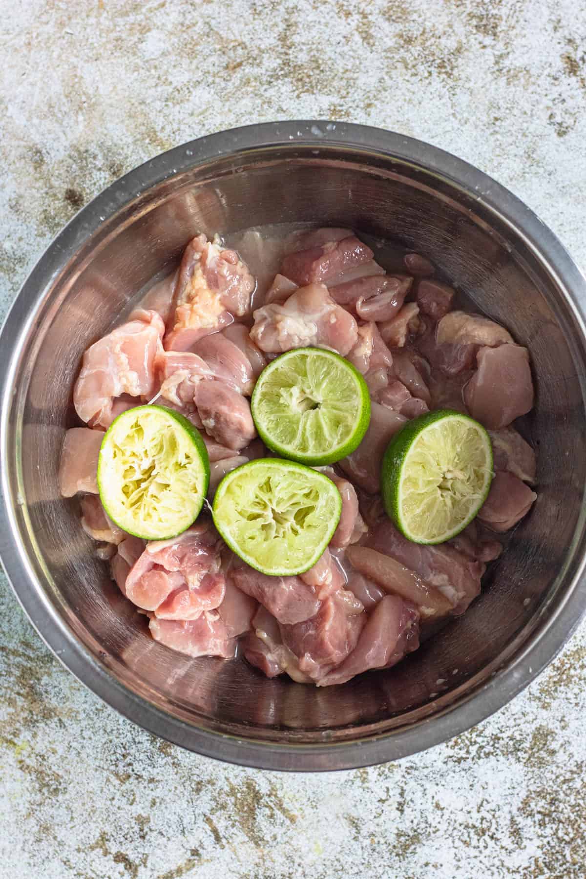 Raw chicken thigh pieces with squeezed limes laying on top in a mixing bowl.