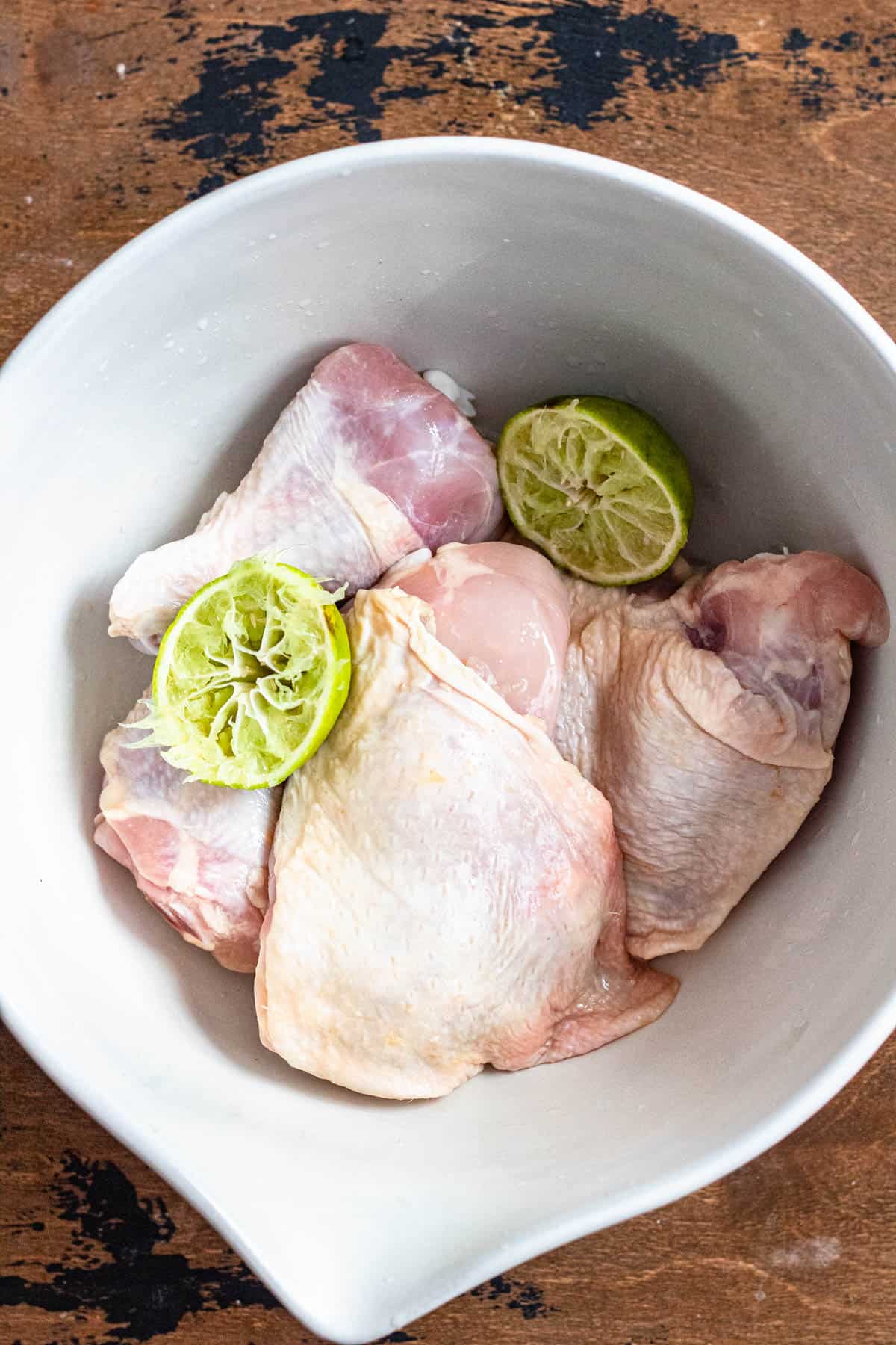 Raw bone in chicken in a mixing bowl with squeeze limes laying on top. 