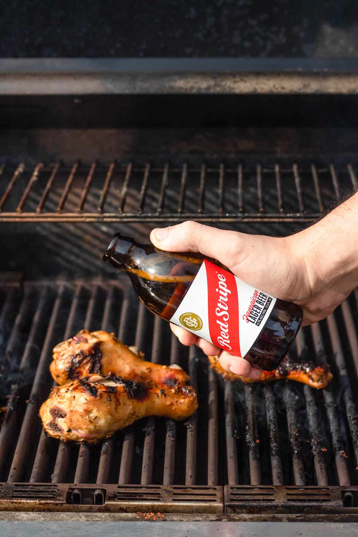 Hand pouring red stripe beer over a marinated and cooking piece of bone-in cihcken on a grill. 