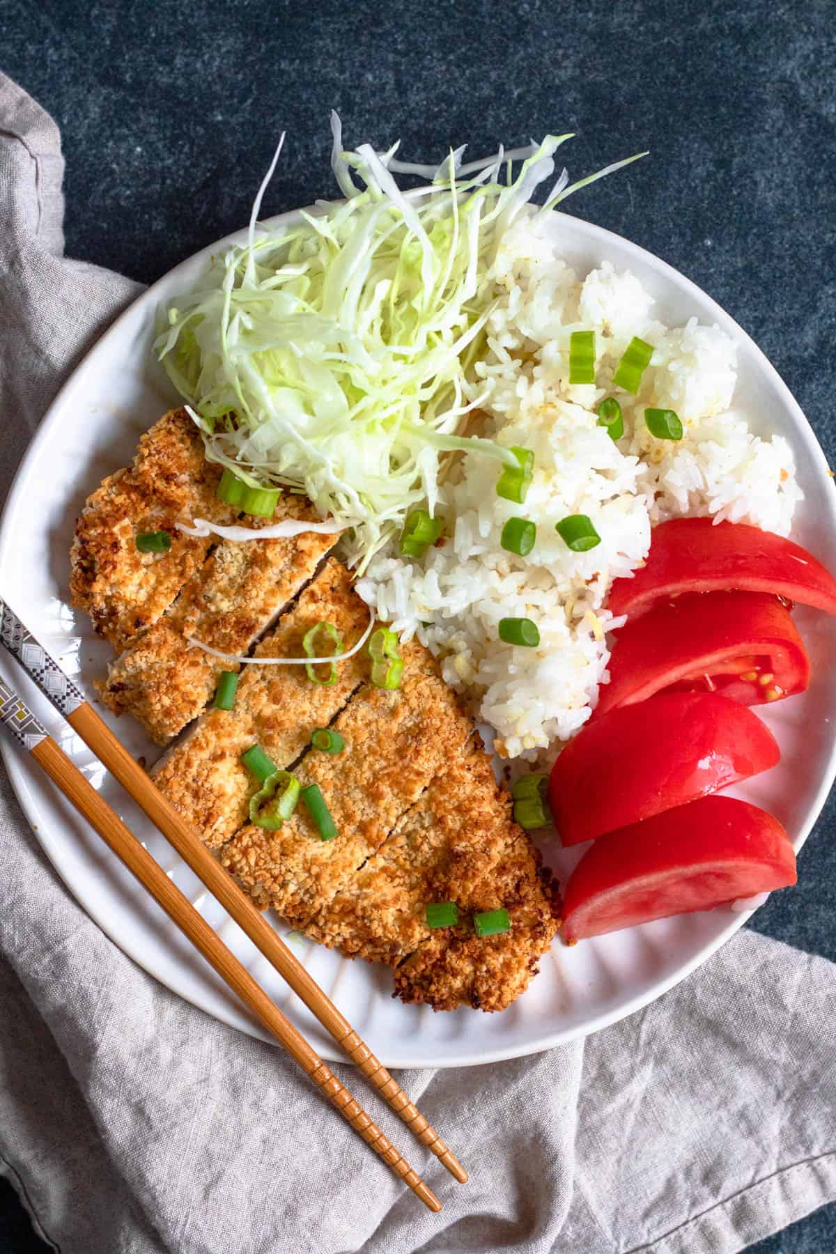 Plate served with chicken katsu, sliced tomatoes, white rice and cabbage with green onions garnished over the top and a side of chopsticks resting on the side. 