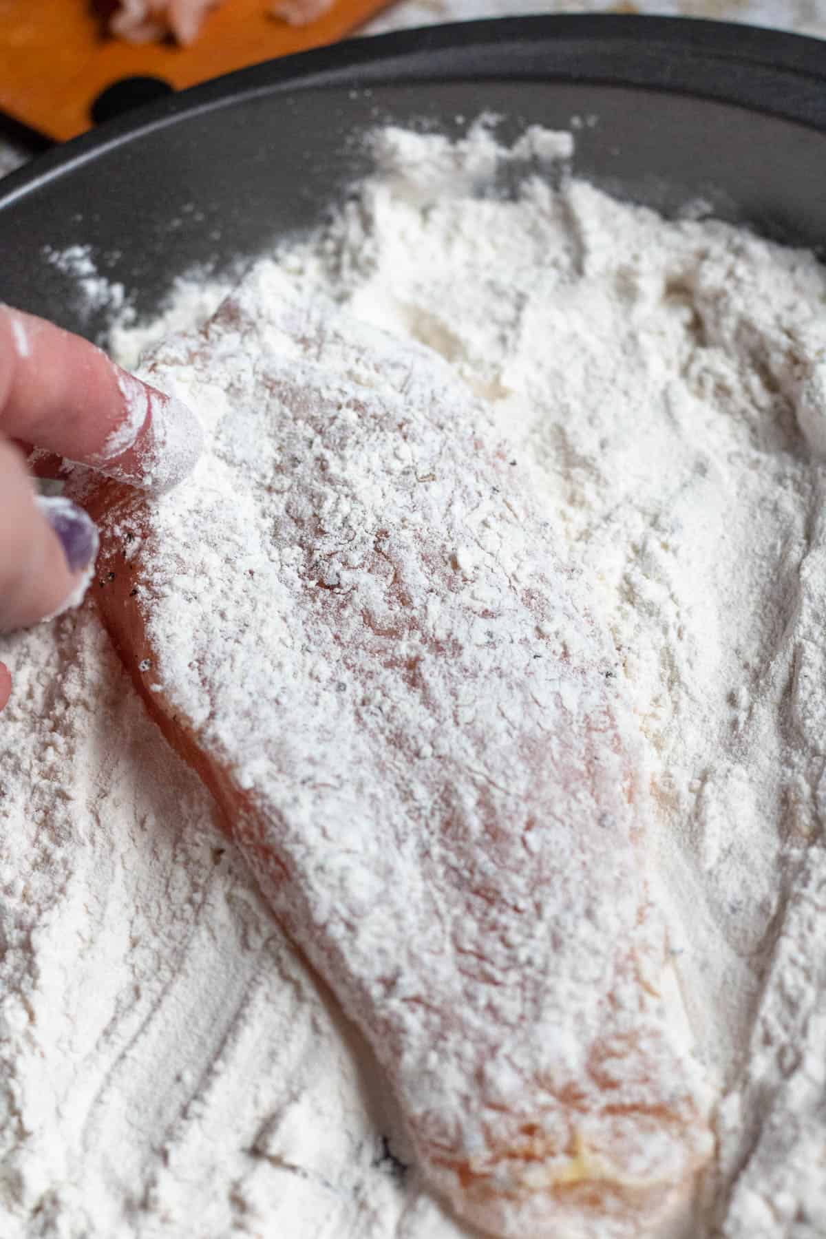 Raw chicken breast being dredged in a pan of flour on both sides. 