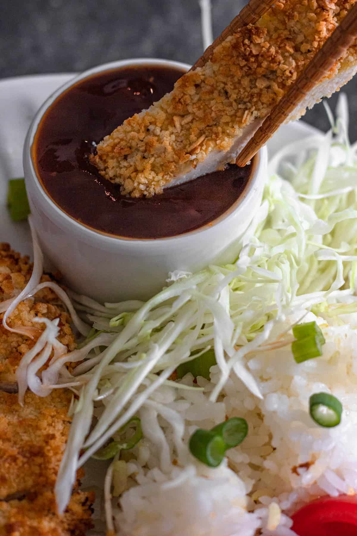 Chopsticks dunking a slice of chicken katsu into a bowl of katsu sauce. 