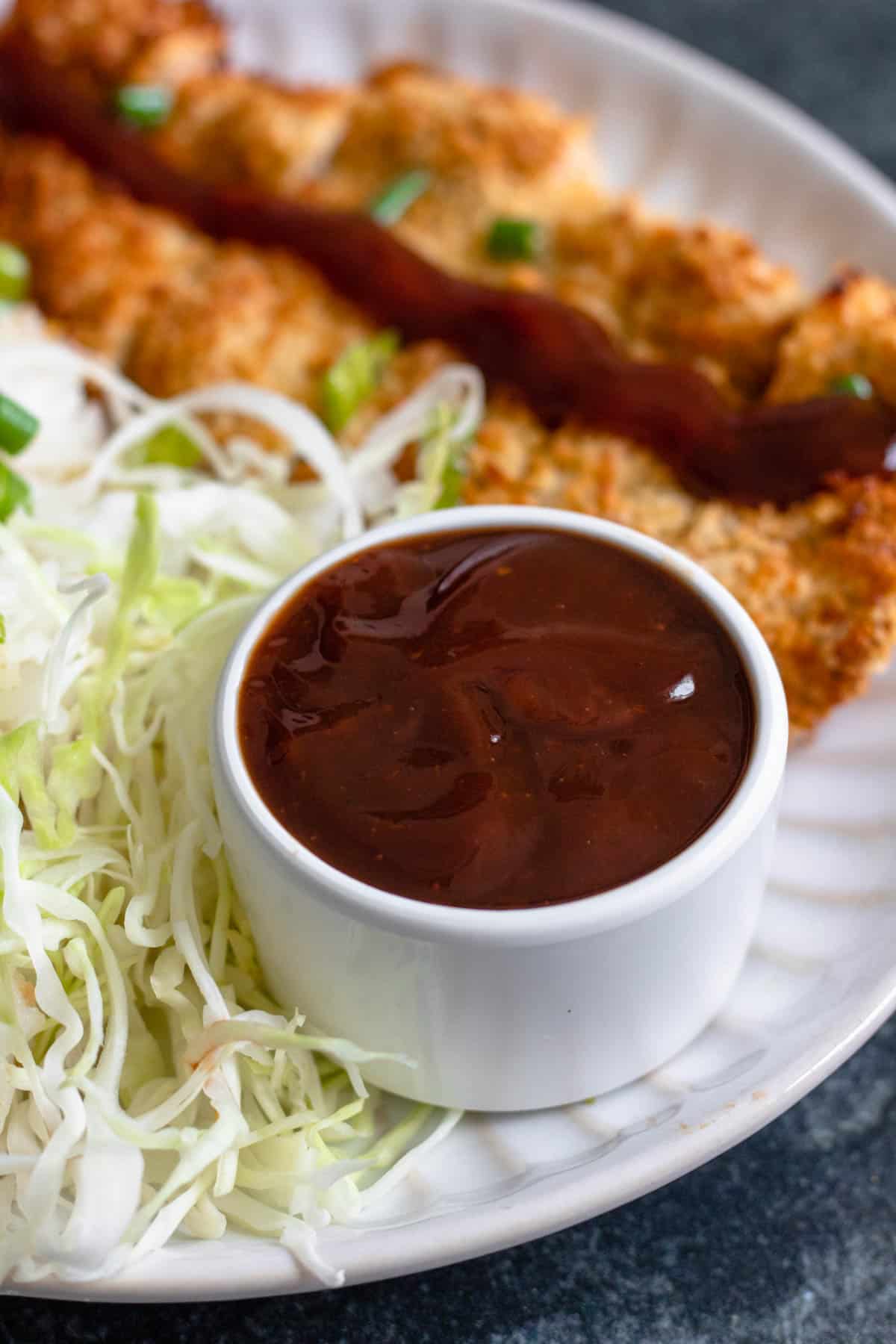 Small bowl of katsu sauce on a plate with chicken katsu and a side of thinly sliced cabbage. 