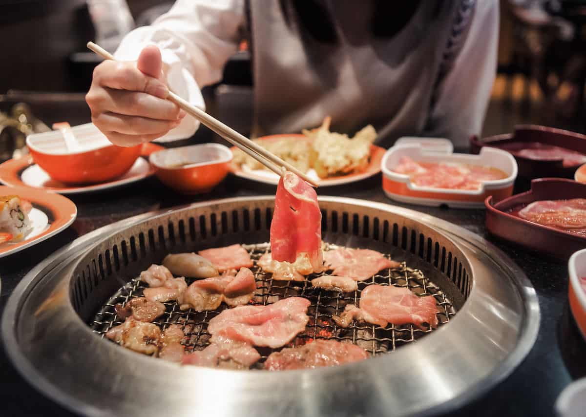 Chopsticks placing a piece of Japanese raw beef onto a tabletop grill with other meats. 