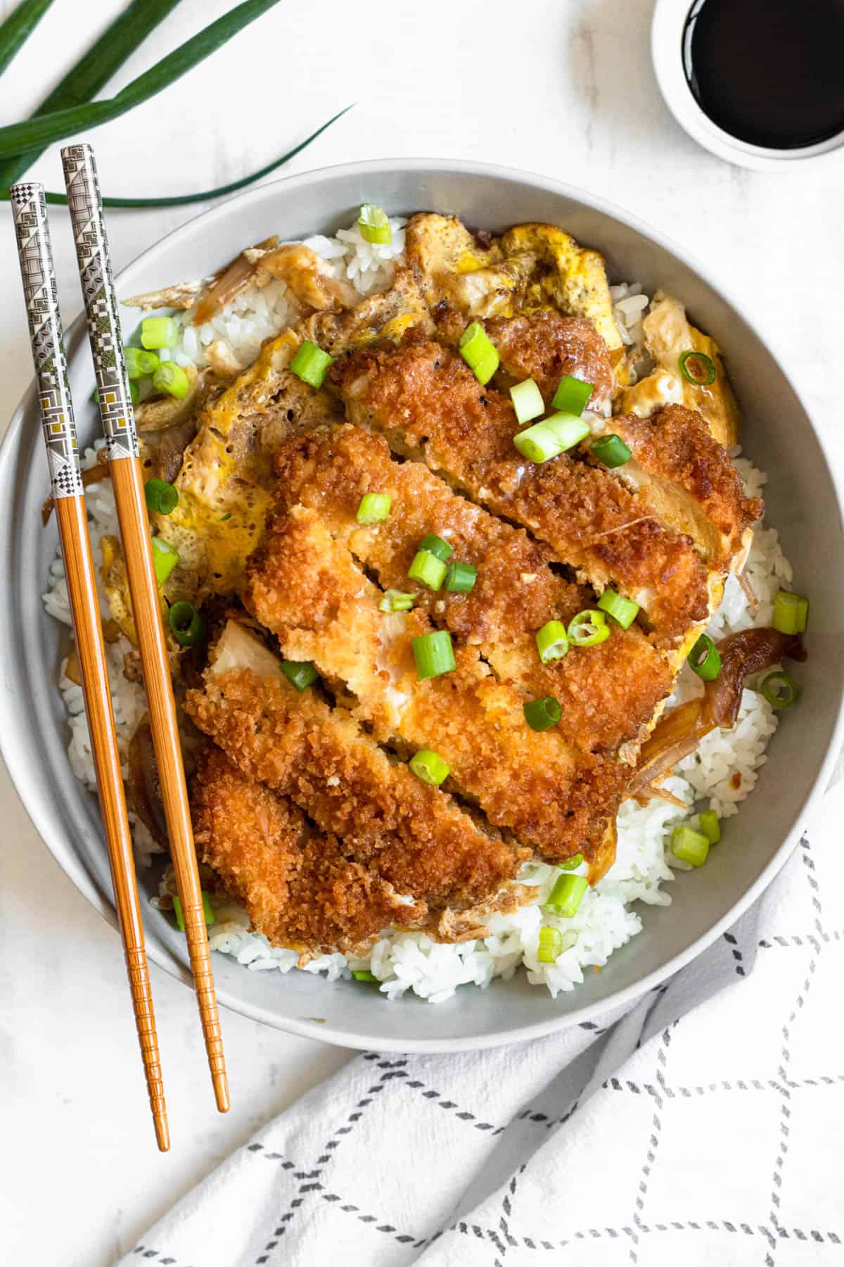 Chopsticks on the side of a bowl of Chicken Katsudon with a bed of white rice. 