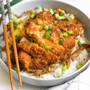 Chicken katsudon over rice and garnished with green onions, sitting next to chopsticks.
