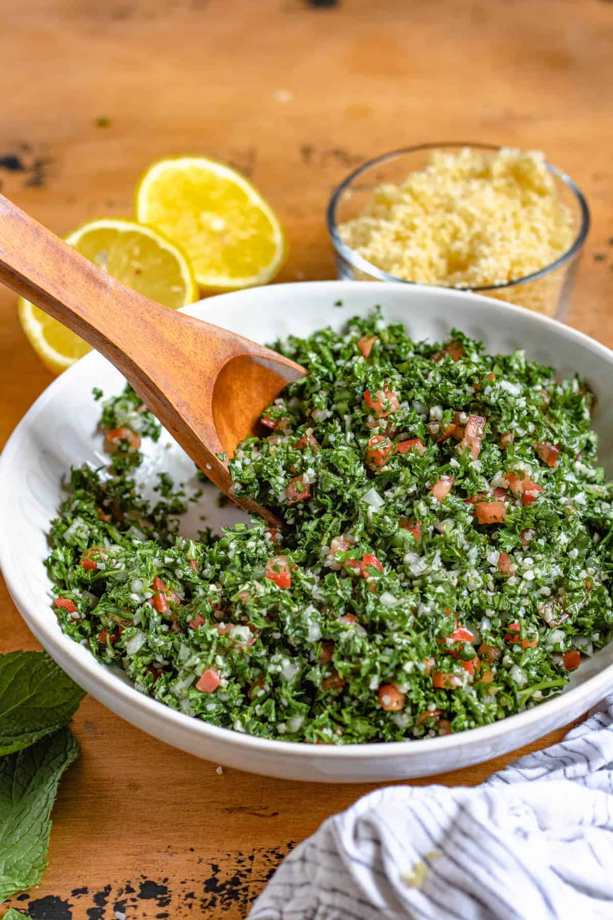 Tabbouli salad in a bowl with a wooden spoon resting in it and lemon halves laying next to the bowl. 