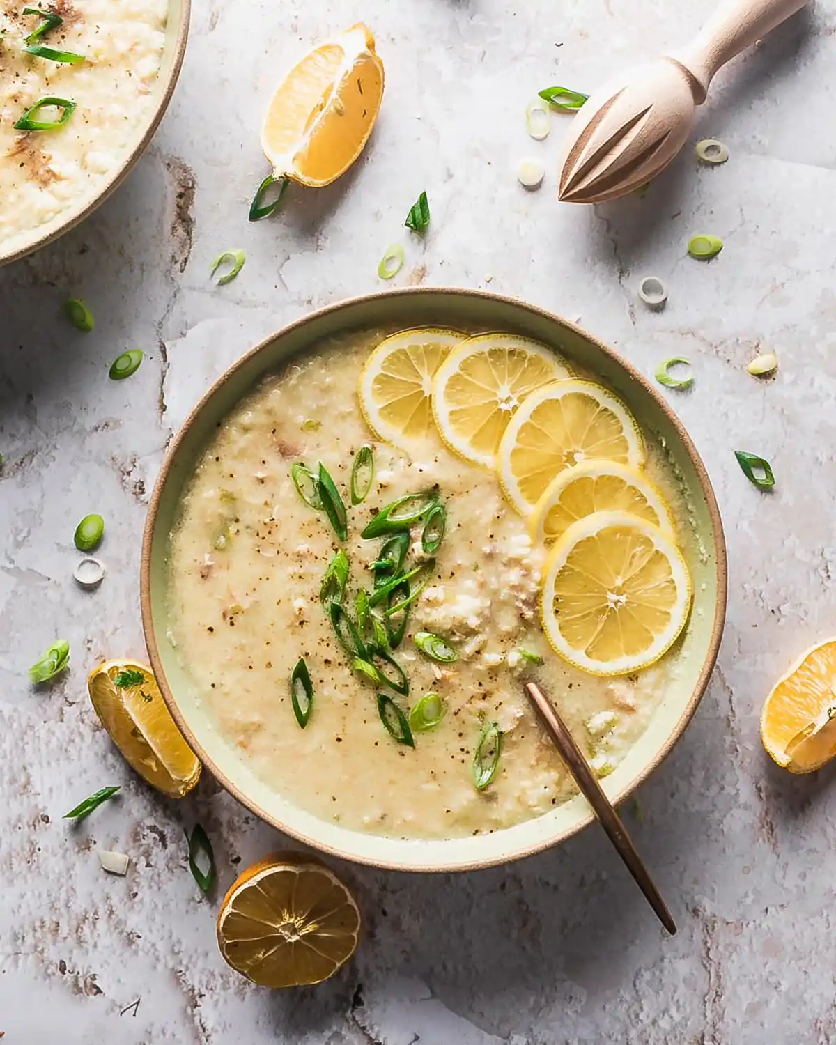 Bowl of greek chicken lemon rice soup with sliced green onions over top and lemon wedges garnished on on the side of the bowl. 