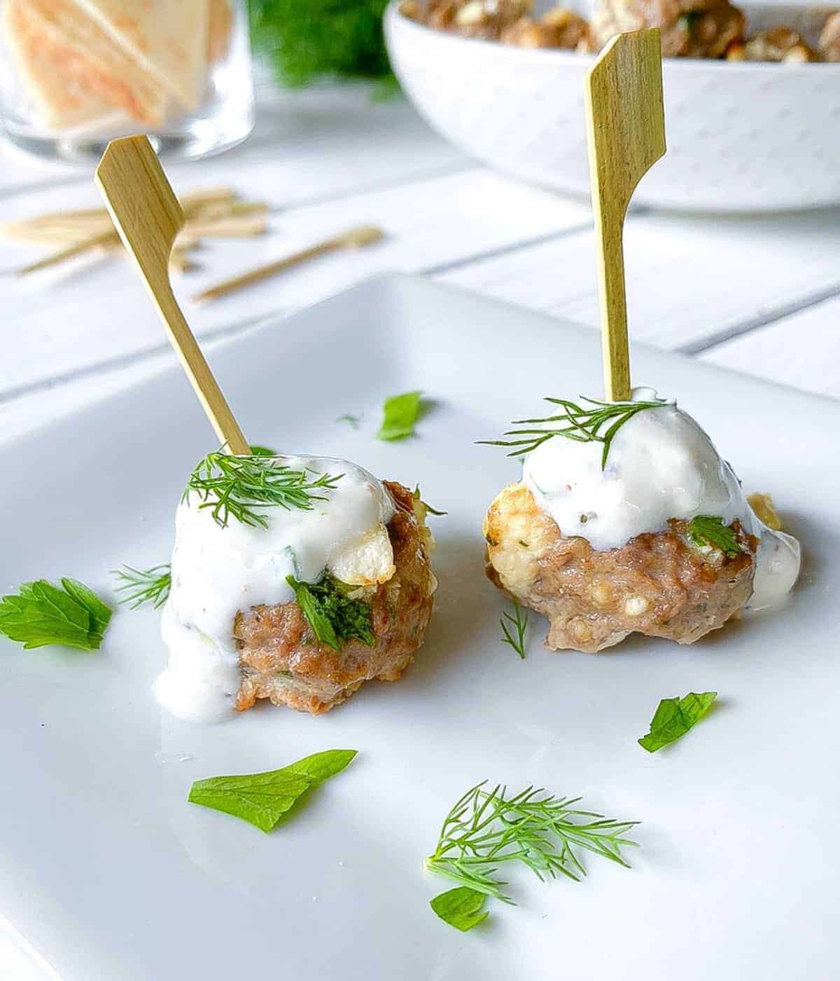 Greek lamb meatballs with a wooden stick coming out of each to use to serve for appetizers, topped with a sacue and fresh dill. 