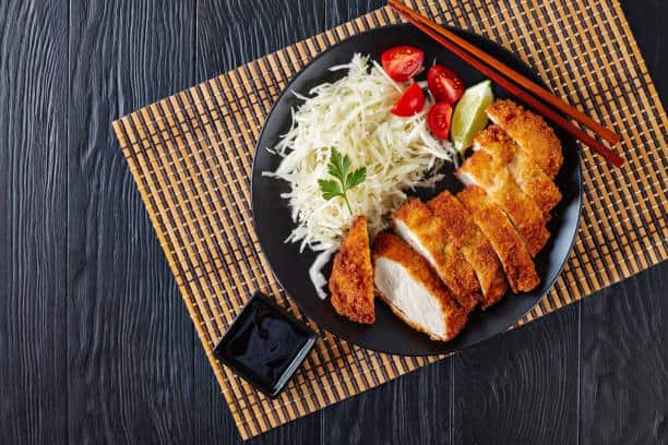 Tonkatsu on a plate with a side shredded cabbage, a lime wedge and sliced tomatoes. 