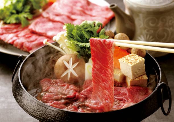 Sukiyaki served in a bowl with chopsticks holding up a pair of thinly sliced beef from the bowl. 