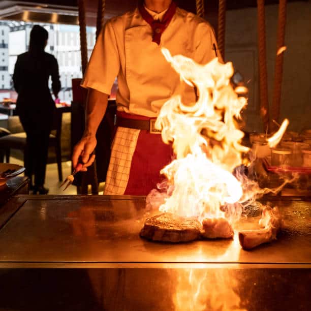 Japanese chef preparing teppanyaki beef on a metal grill top with flames shooting around the steak. 
