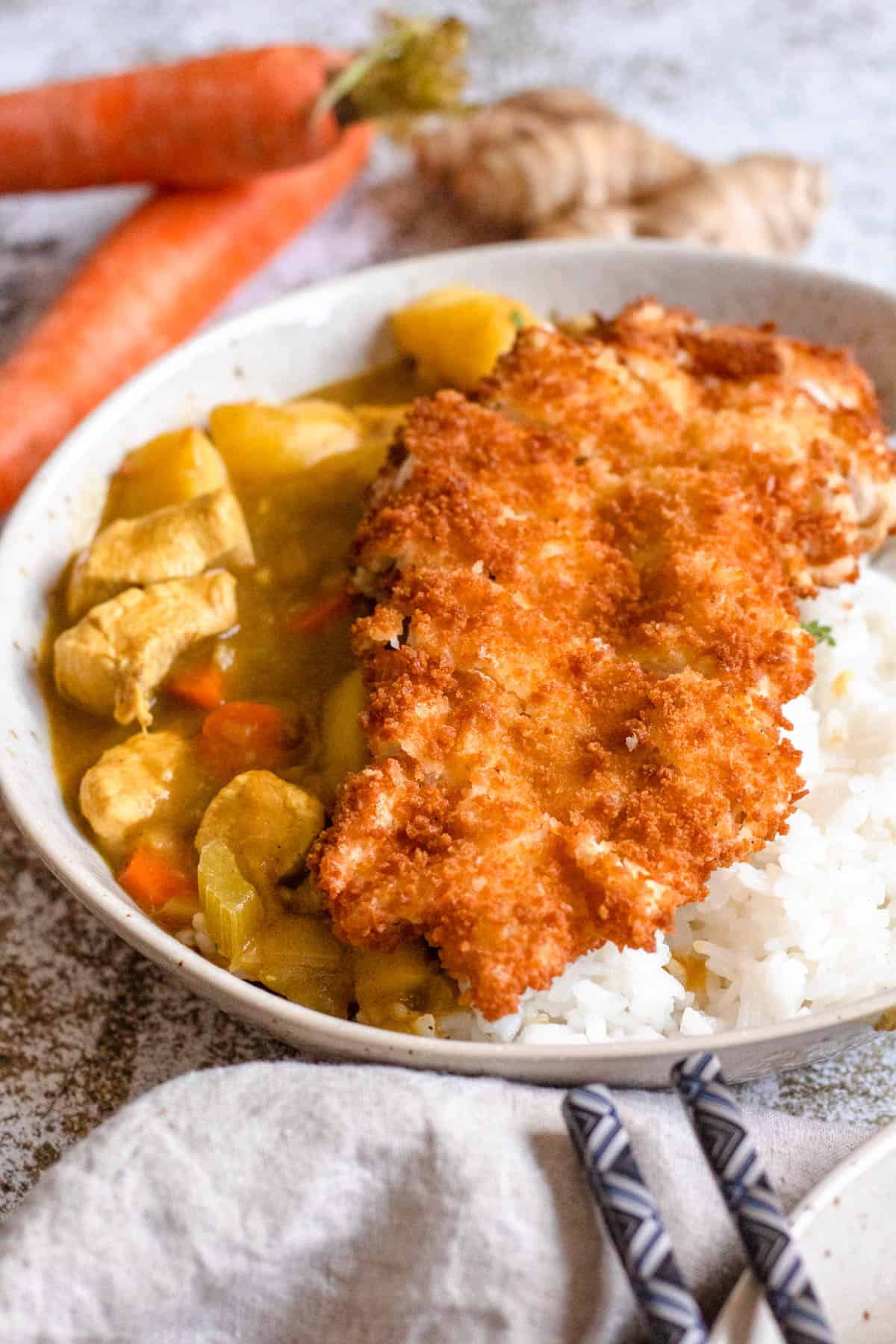 Chicken katsu curry recipe served in a bowl with fresh ginger and whole carrots next to it. 