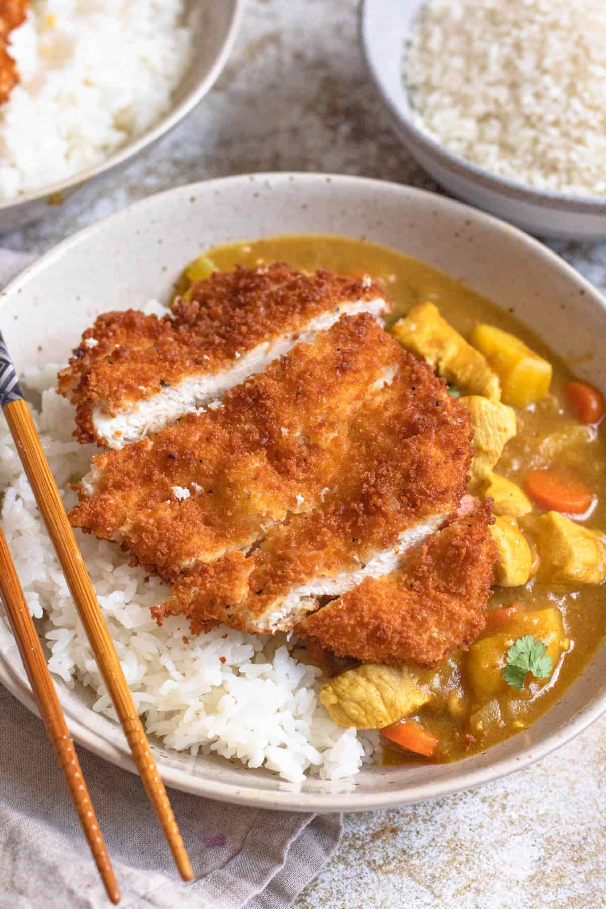 Bowl of white rice with chicken katsu served over top an some chicken curry on the side and a pair of chopsticks on the edge. 
