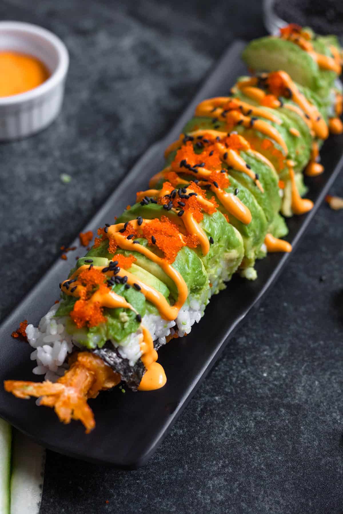 Dragon roll sushi recipe served on a rectangular serving dish drizzled with sriacha mayo with a side of dipping sauce. 