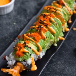 A platter with dragon roll sushi on it topped with avocado, spicy mayo, masago, and black sesame seeds.
