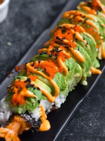 A platter with dragon roll sushi on it topped with avocado, spicy mayo, masago, and black sesame seeds.