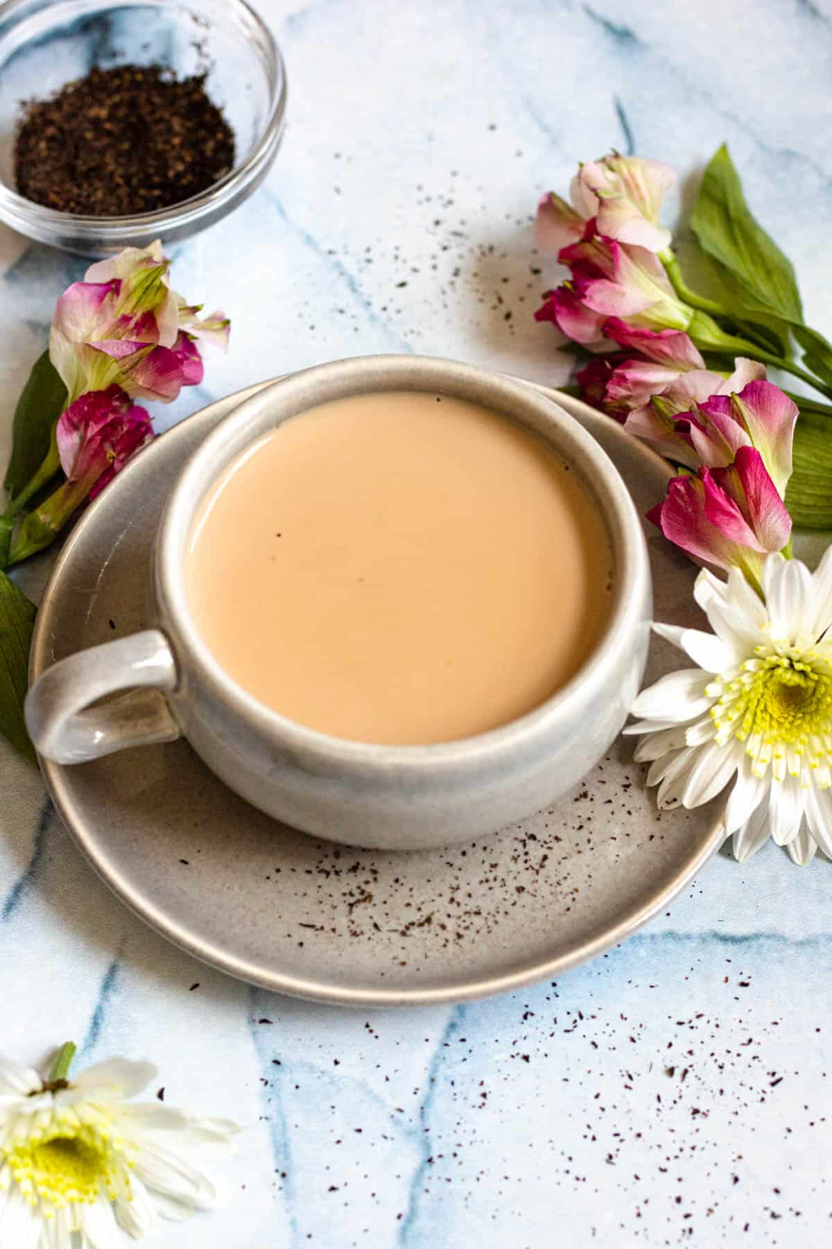Royal milk tea served in a coffee mug served on a saucer with fresh flowers laying around it. 