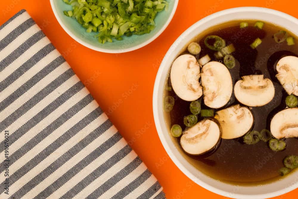 A bowl of Japanese clear soup with green onions and mushrooms and a bowl of sliced green onions next to it. 