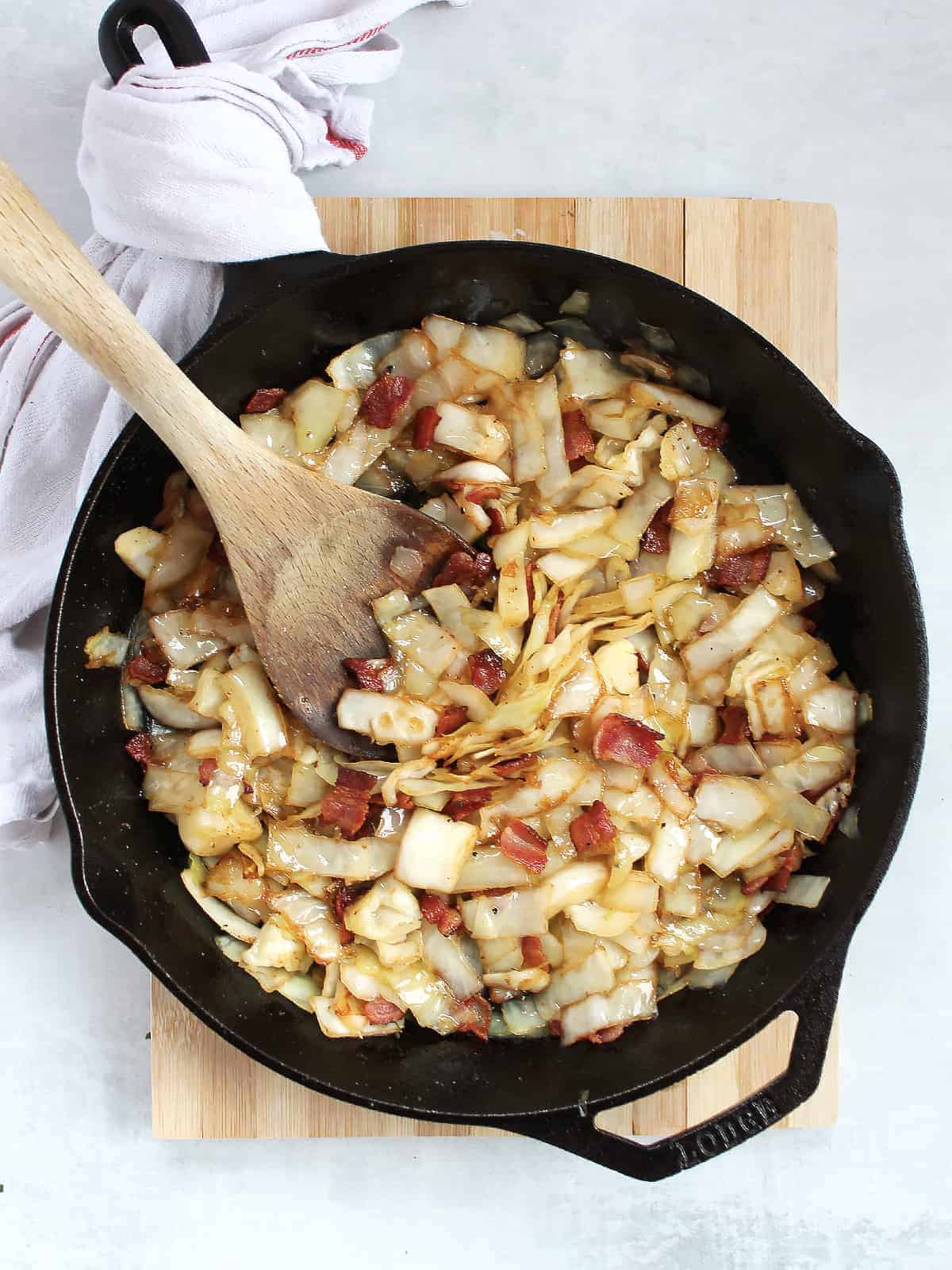 German fried cabbage cooked with bacon in a cast iron pan and a wooden spoon resting in it. 