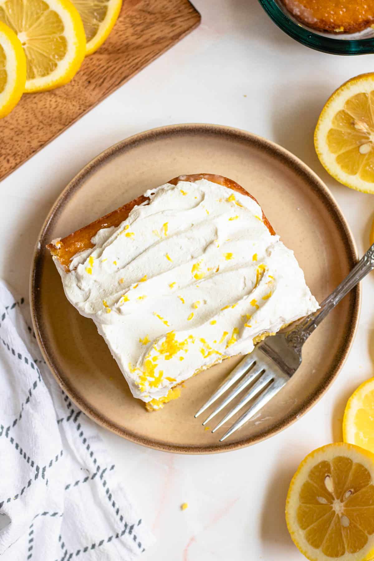 Slice of lemon cake with lemon zest over the top on a plate with a fork resting on the side. 