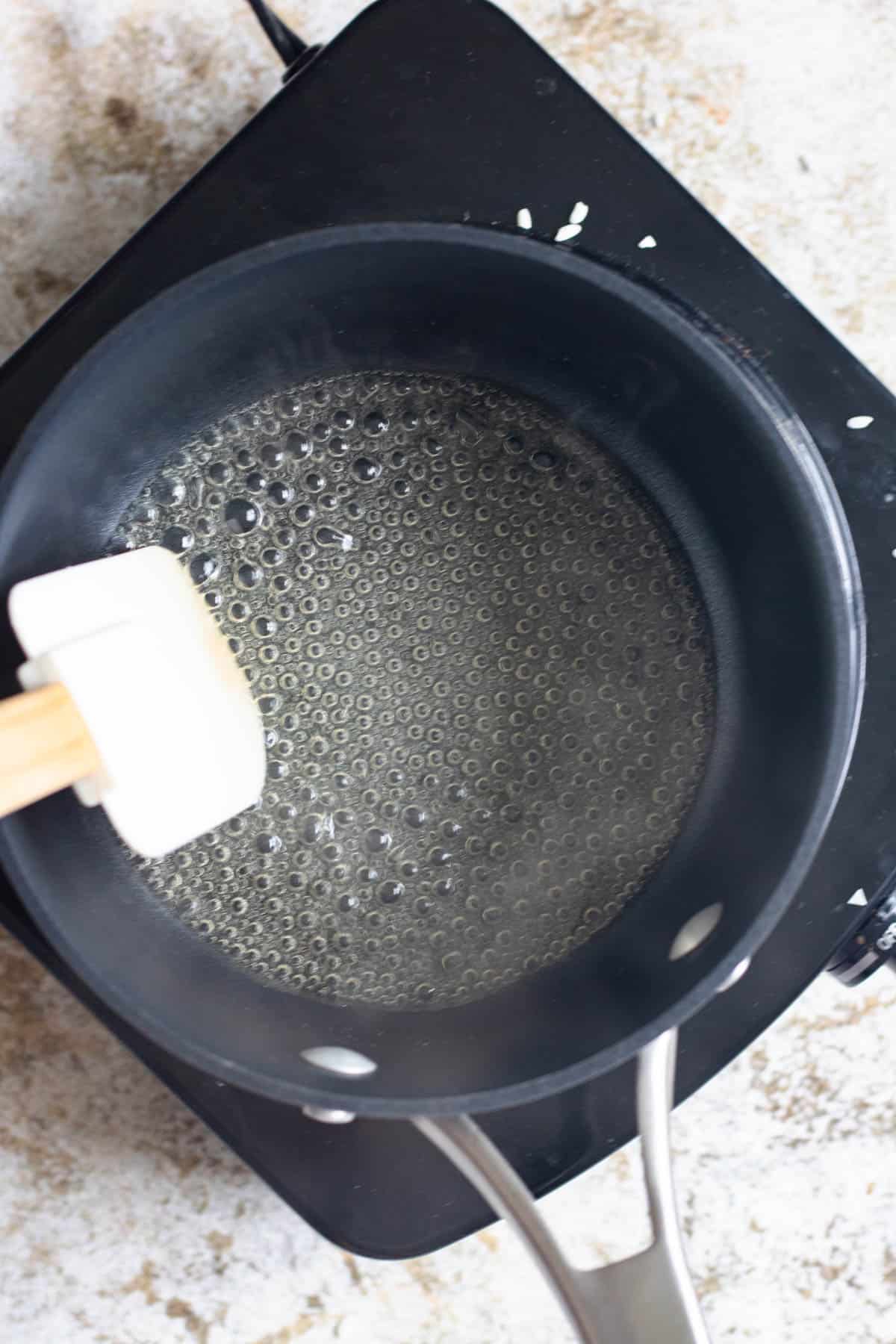 Sugar mixture coming to a boil in a saucepan with a spatula stirring it. 