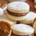 Pinterest image of a close-up shot of Alfajores Argentinos lying stacked and next to each other, with one filled with dulce de leche but not sandwiched with another cookie.
