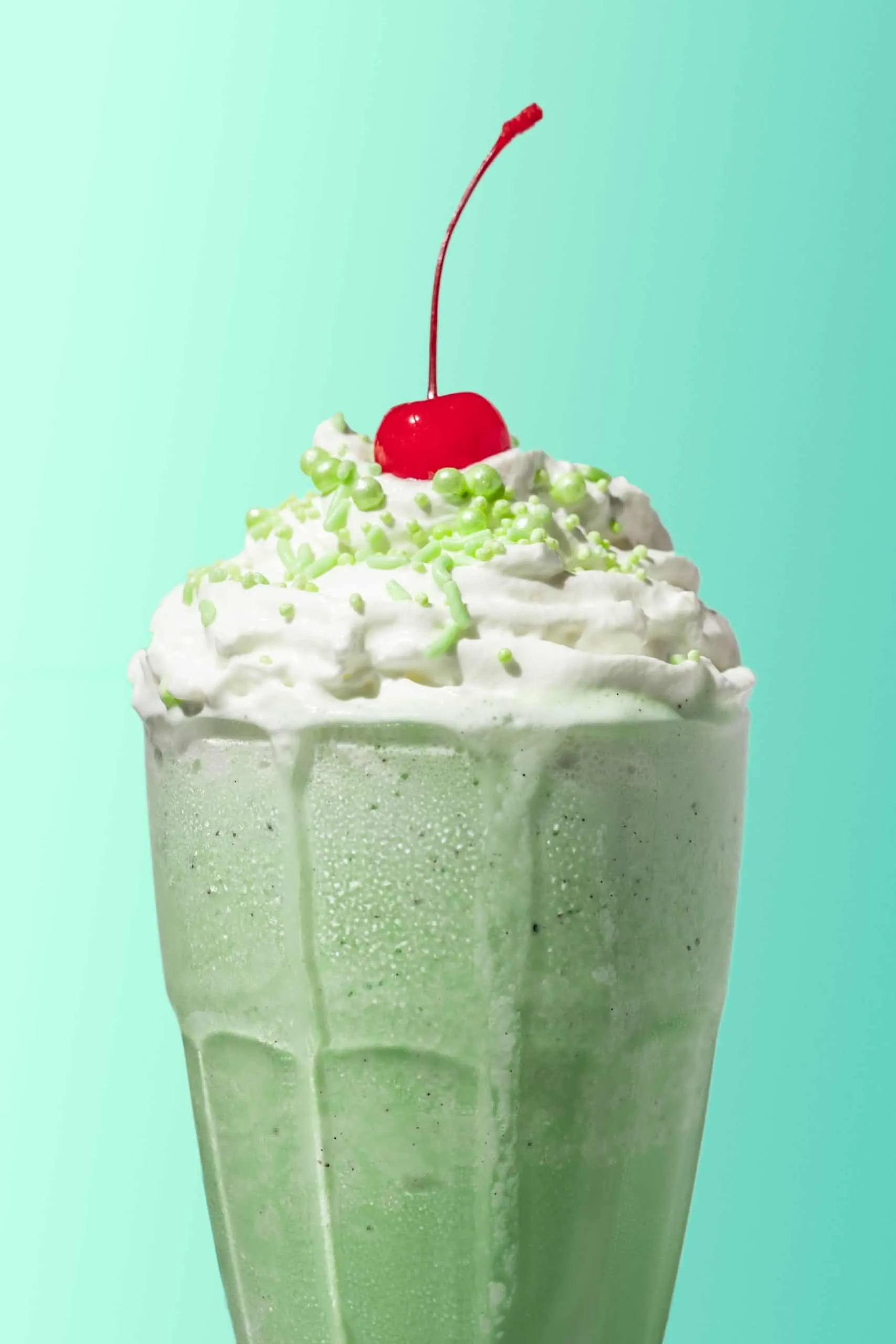 Boozy shamrock shakes in a shake glass with whipped cream and a maraschino cherry on top. 