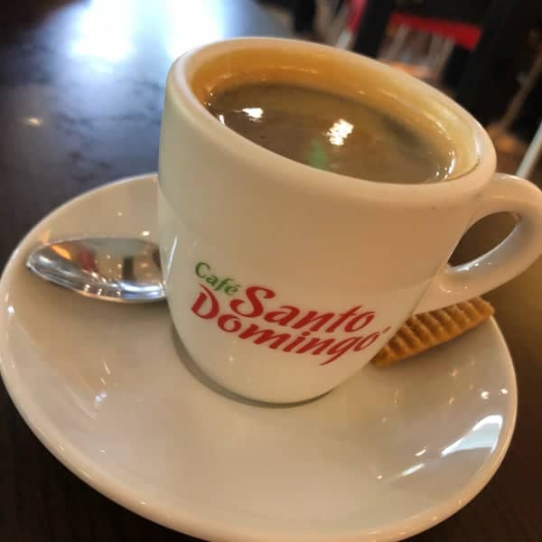 A coffee mug with the text Care Santo Domingo on a saucer and a spoon resting on it. 