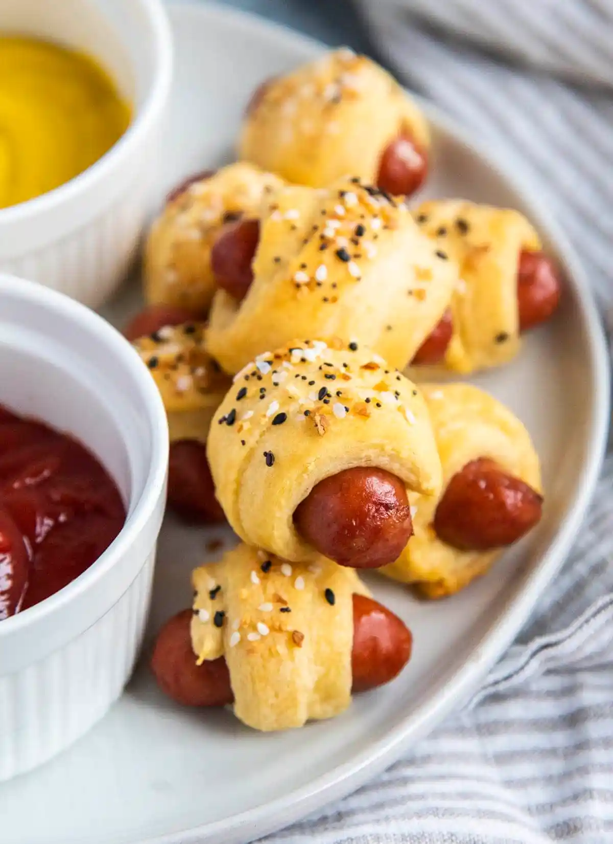 Everything bagel pigs in a blanket on a plate with a side of dipping sauce. 