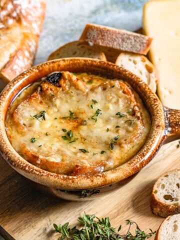 A crock of Instant Pot French Onion Soup topped with melted cheese, sitting next to fresh herbs and sliced baguette.