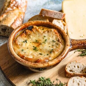 A crock of french onion soup on a cutting board, surrounded by fresh herbs and baguettes.