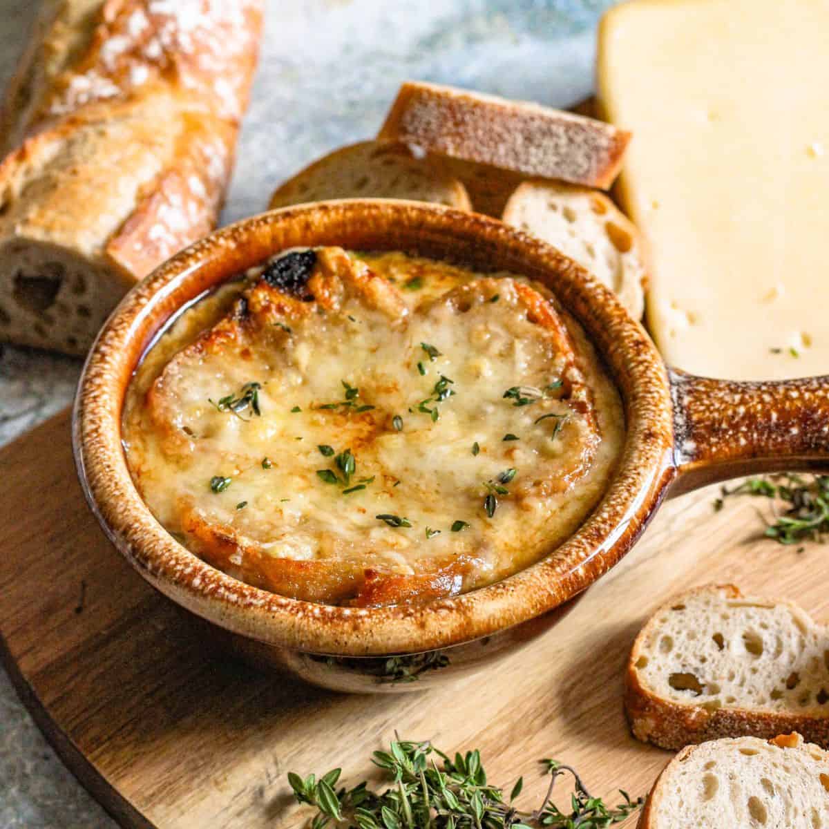 A crock of french onion soup on a cutting board, surrounded by fresh herbs and baguettes.