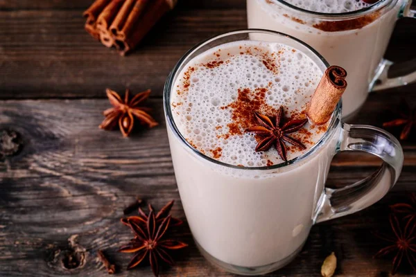 Two glass mugs of chai lattes with a cinnamon stick added to it along with a star anise. 