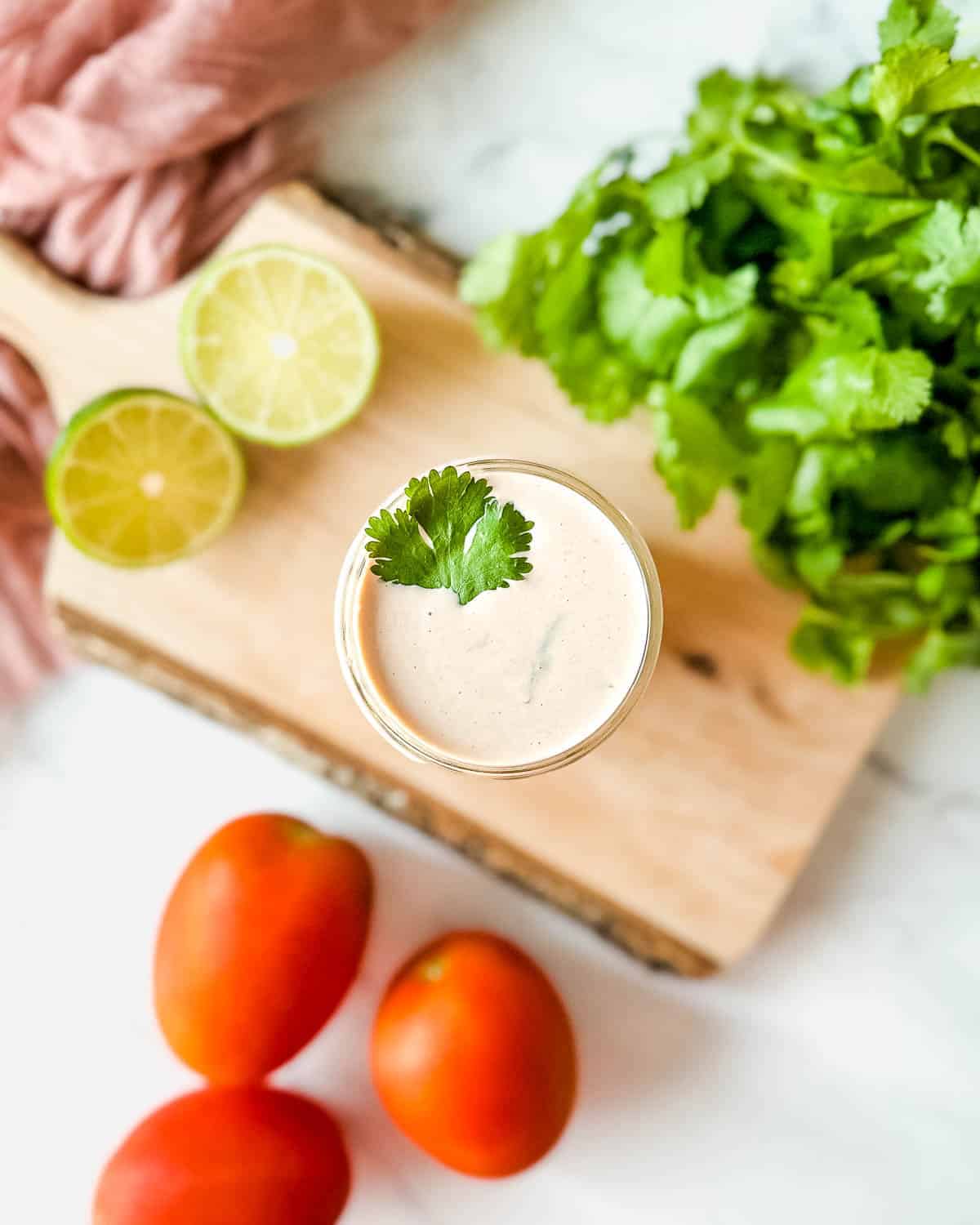Creamy salsa dressing in a small glass jar with a cilantro leaf garnished on top and lime wedges, cilantro and tomatoes laying next to it. 