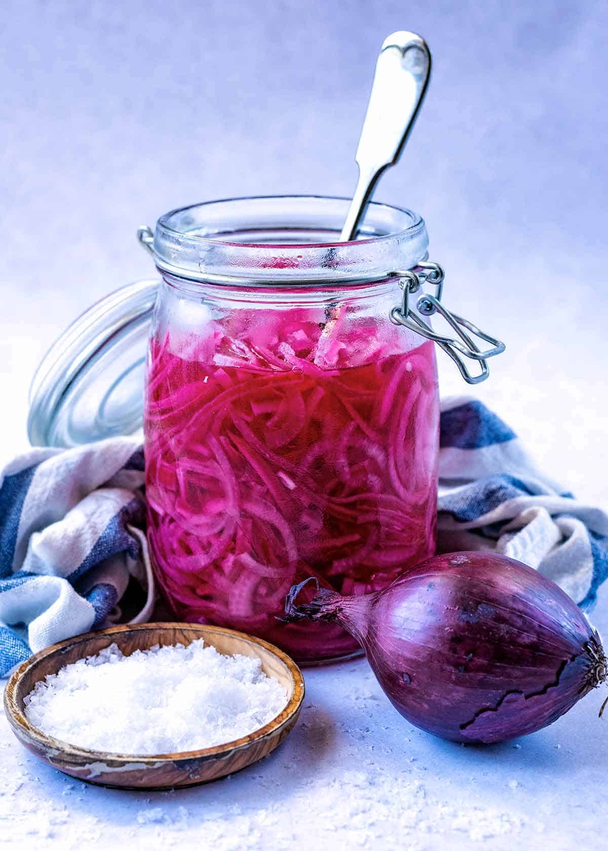Whole red onions and salt in a bowl next to a jar of quick pickled red onions. 
