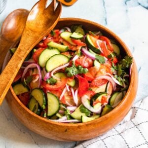 Tomato and cucumber salad in a wooden bowl with wooden serving utensils next to it.
