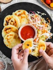 Hand pulling apart a pupusa, in front of a plate of pupusas, curtido, and dipping sauce.