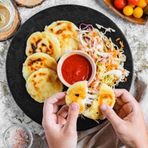 Hand pulling apart a pupusa, in front of a plate of pupusas, curtido, and dipping sauce.