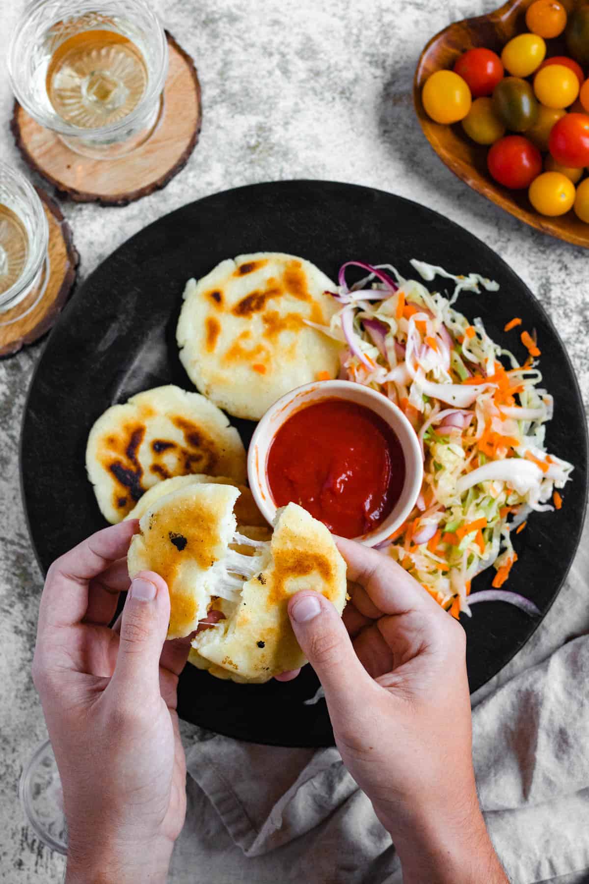 Hand holding a freshly made pupusa over a platter of them with curtido on the side, breaking it apart to show the melty cheese inside.