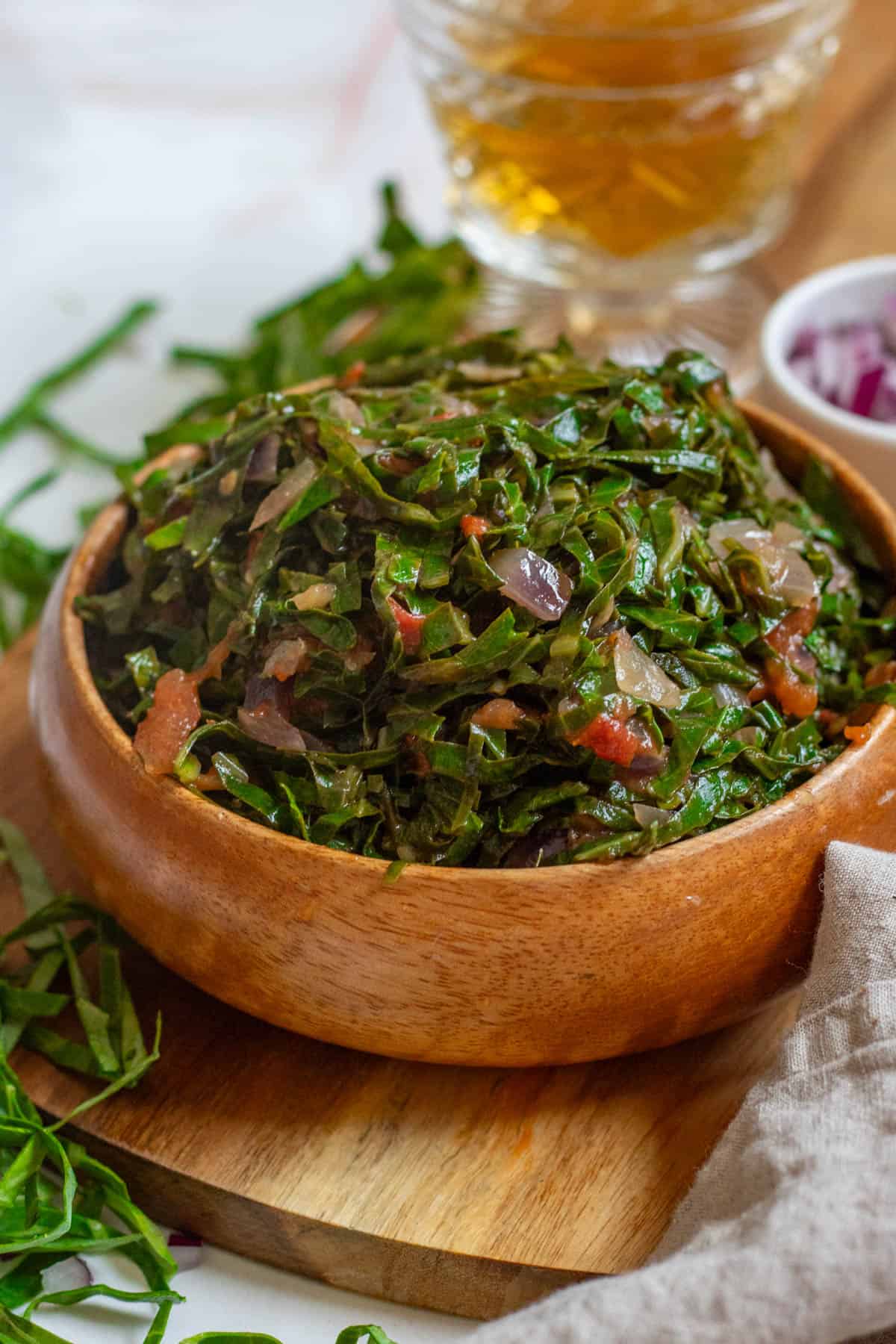 A wooden bowl filled with Sukuma Wiki with red onions in the background. 