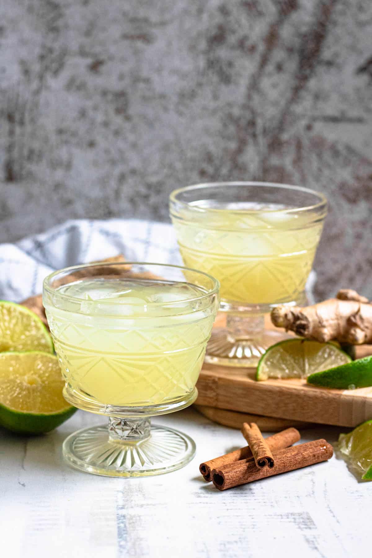 Two small glasses with ice and homemade ginger juice served in it with cinnamon sticks and lime wedges next to them. 