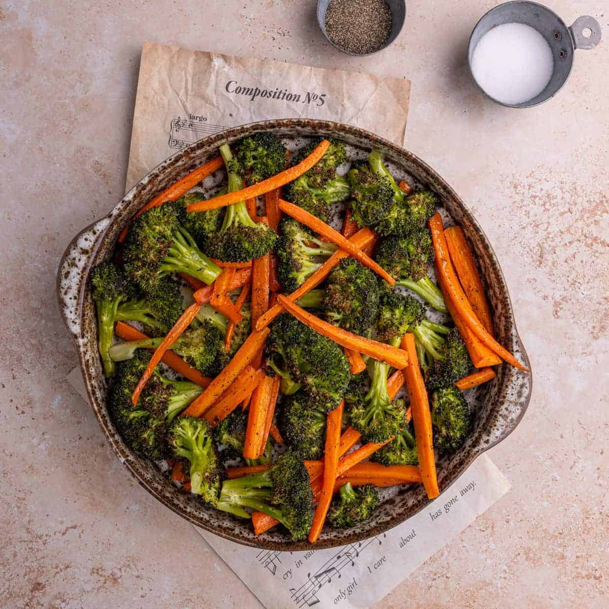 Roasted broccoli and carrots served in a round bowl with small bowls with salt and pepper. 