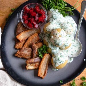 A plate of stegt flaesk with cooked pork belly, potatoes, and a cream parsley sauce with a spoon and pickled beets on the side of the plate.