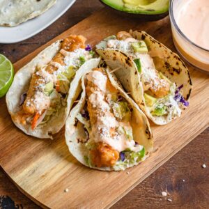 Fish tacos (tacos de pescado) on a wooden cutting board, drizzled with sauce, with lime and avocado on the side.