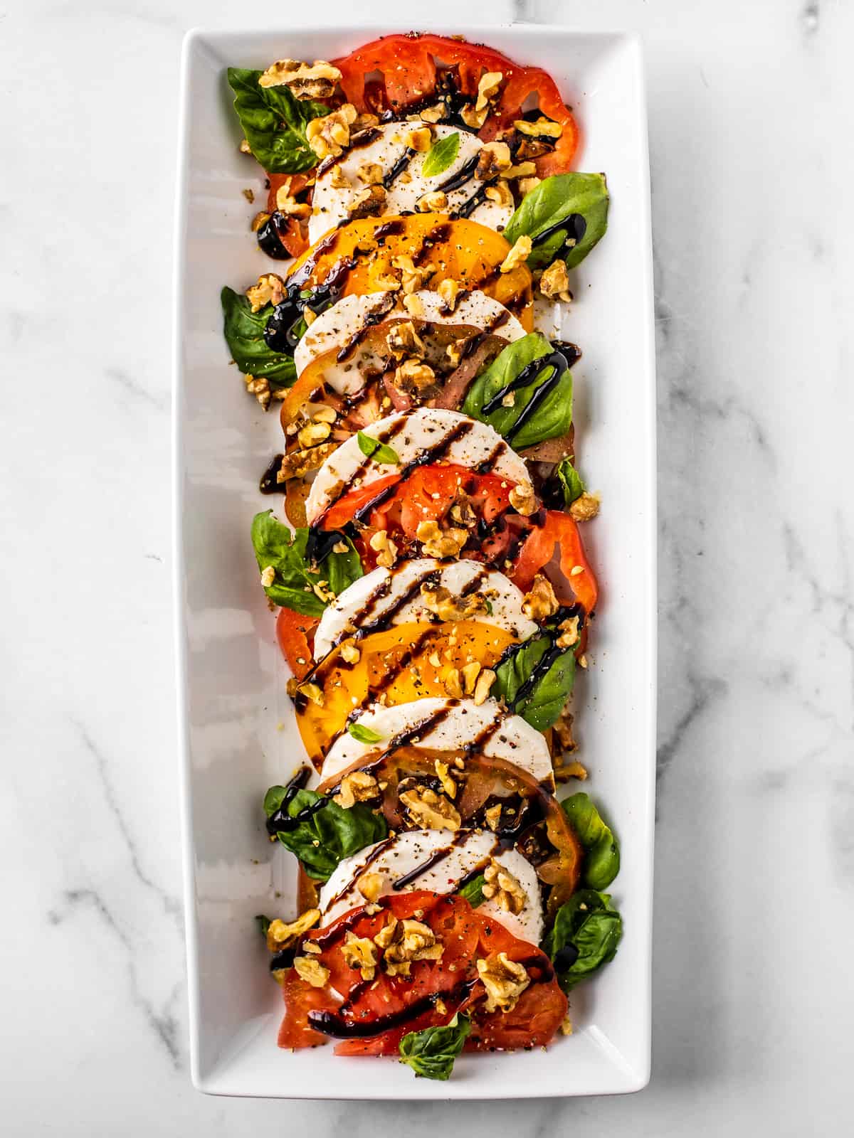 Caprese salad served on a white rectangular platter drizzled with balsamic reduction and chopped walnuts. 