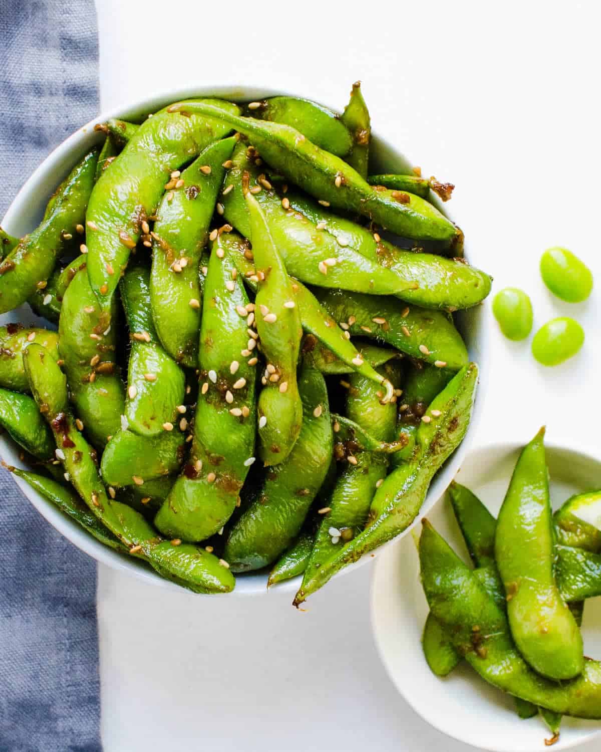 Spicy edamame in a bowl with sesame seeds garnished on top and a couple edamame pieces served in a small bowl. 
