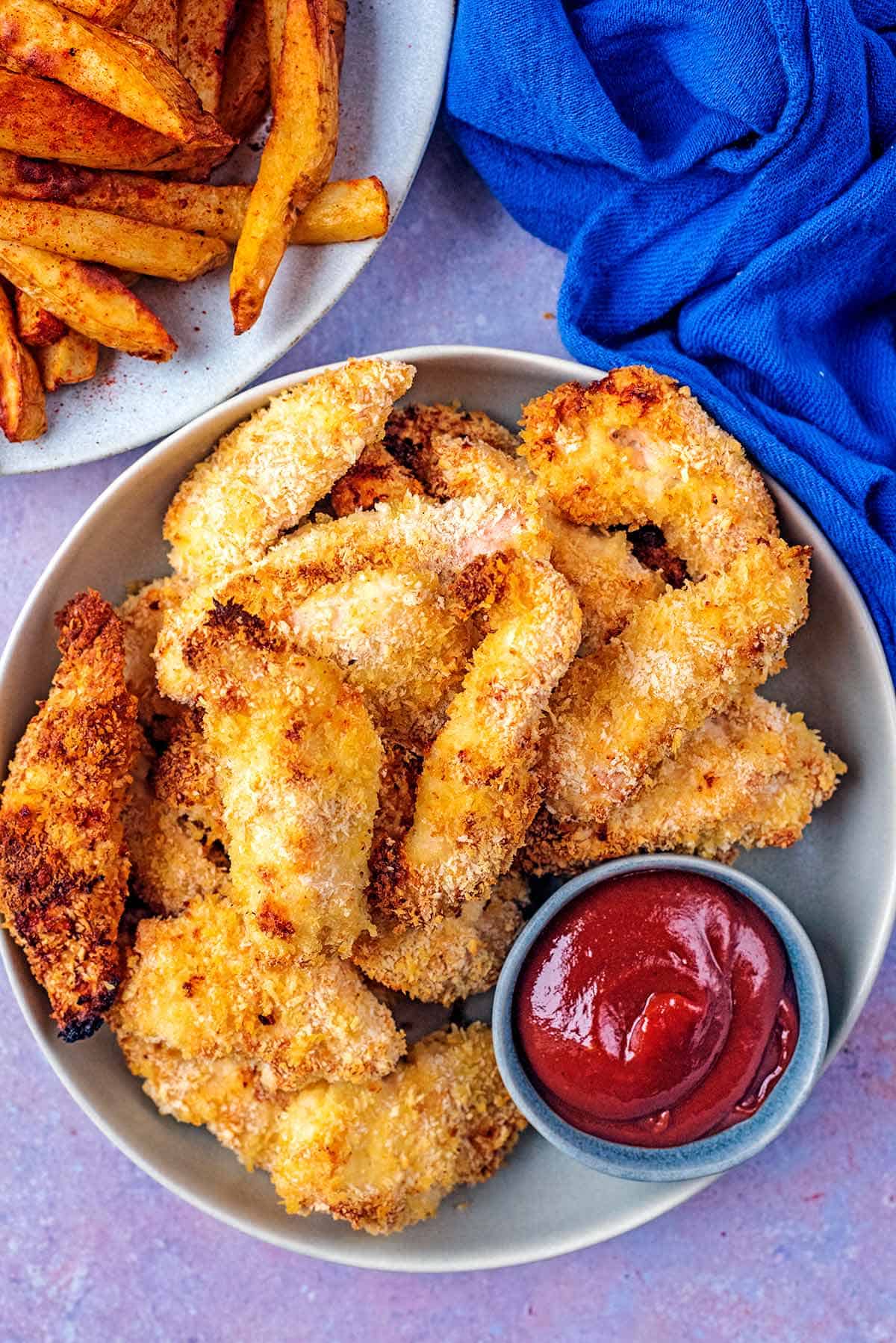 Plateful of crispy chicken goujons with a side of ketchup on the plate and a plate of french fries served next to it. 