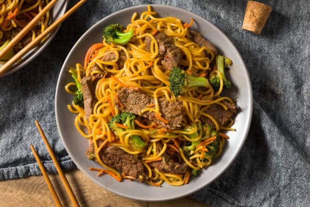 Beef lo mein served on a plate with stir fried broccoli and carrot matchsticks. 