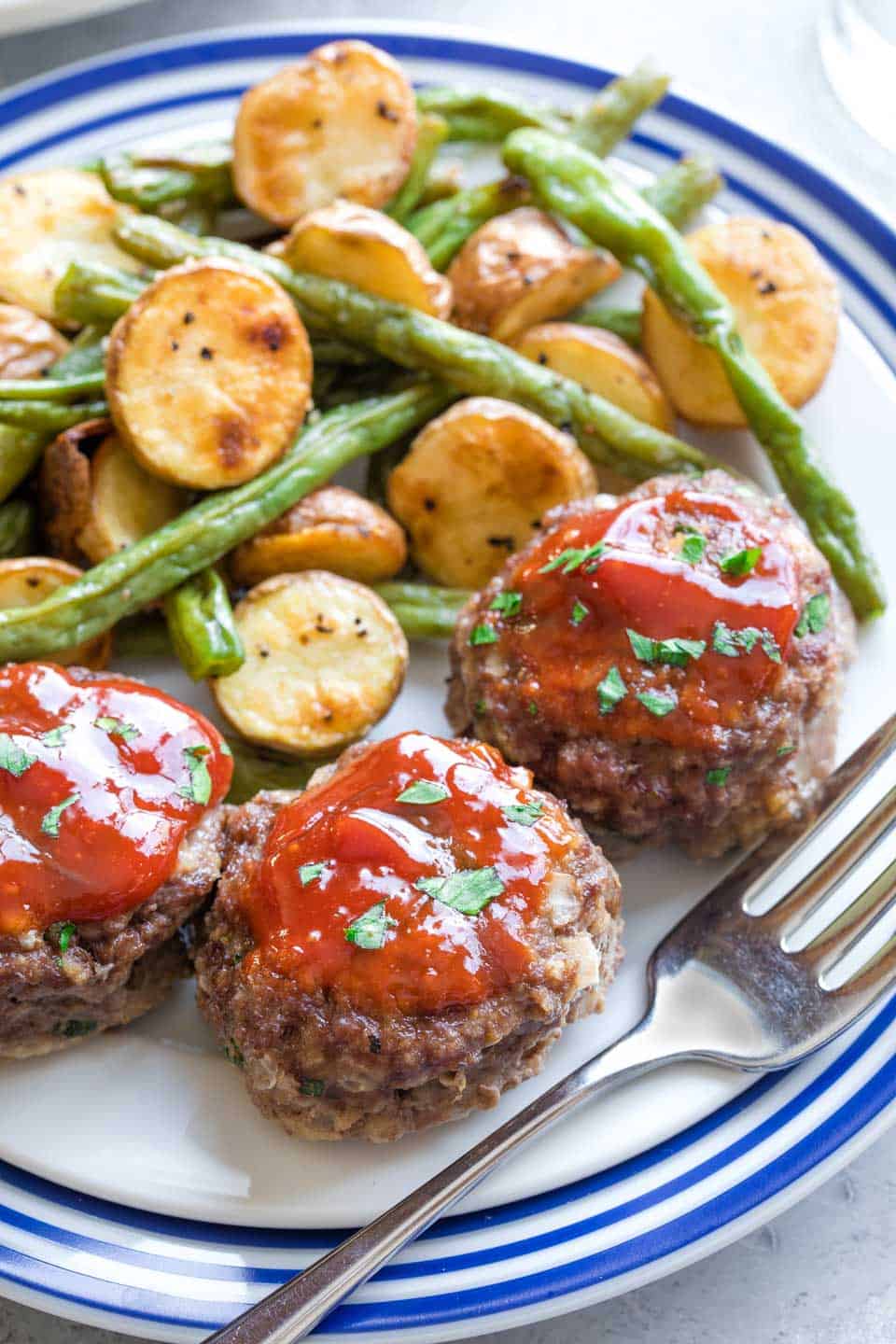 Meatloaf muffins with a red sauce served over the top on a plate that has potatoes and green beans served with it. 